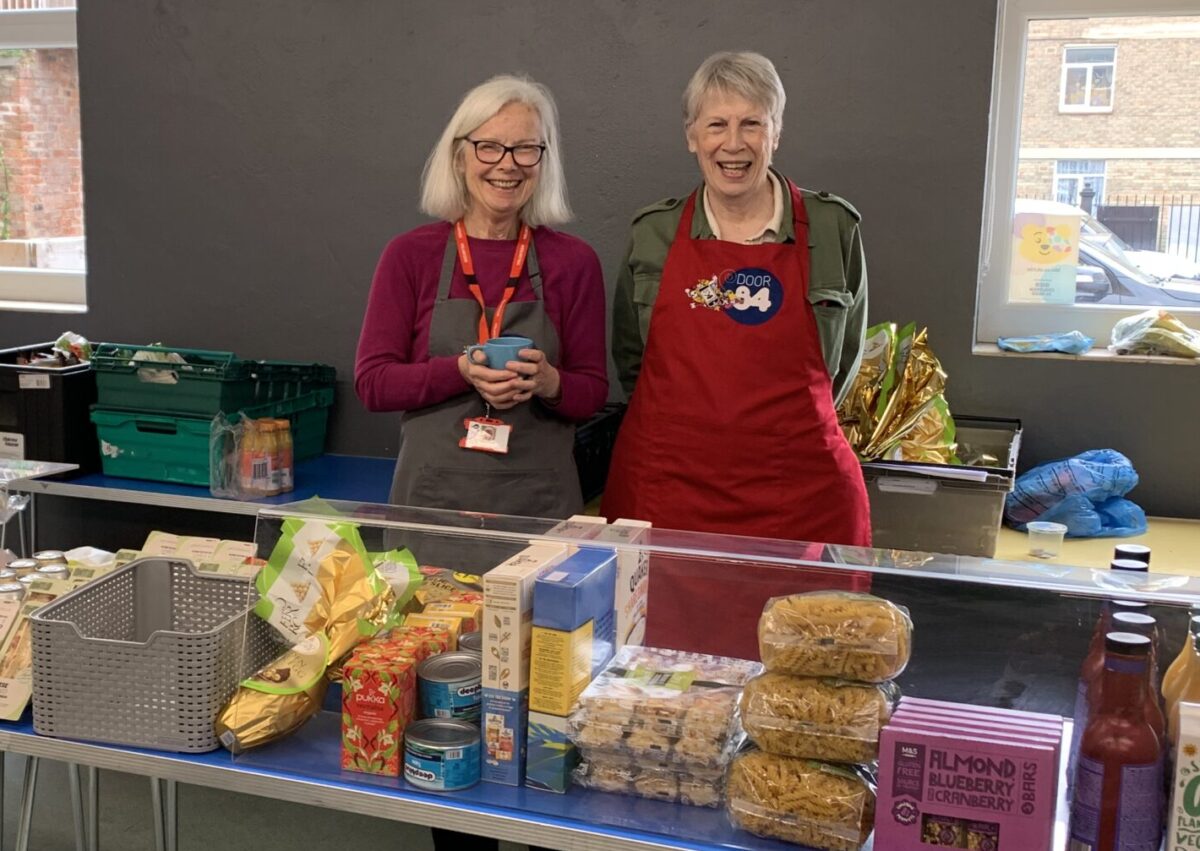 two food bank volunteers behind a table of food, smiling.