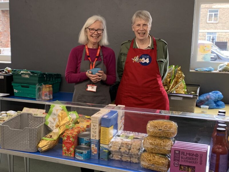 two food bank volunteers behind a table of food, smiling.